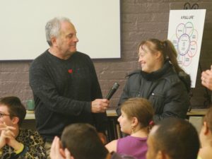 Man holds out microphone to young woman in front of small audience.