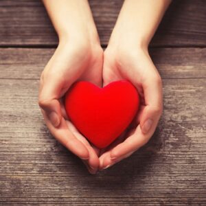 Two light-skinned hands holding a red heart against a dark grain wood background