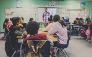 A teacher in front of their classroom, photographed from the students' perspective.