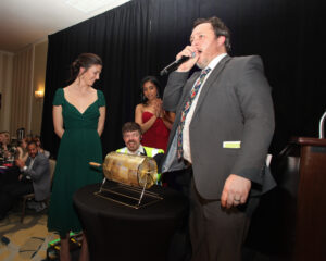 Preparing for the Golden Ticket raffle drawing. A woman in green stands on the left, next to the golden raffle drum and a woman in red. A man in a gray suit speaks into a microphone as another man pulls the winning ticket.