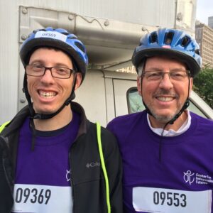 Caleb and his father smile at the camera, both wearing purple shirts and blue helmets