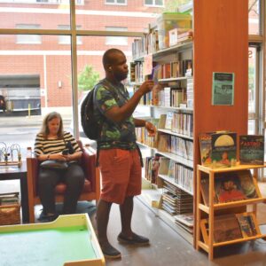 Young man taking a book off of a shelf at the library with many books surrounding him and glass windows in the background.