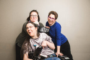 Sarah smiles in foreground of photo with two women standing behind her, one in green and the other in blue.