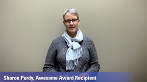 A woman stands in front of a gray/green background with a gray shirt and  light blue scarf on. The purple intro banner reads "Sharon Purdy, Awesome Award Recipient"